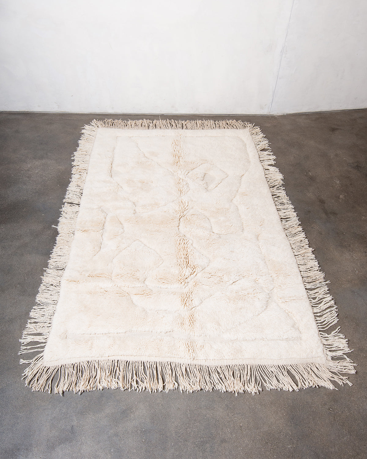 Berberlin x Malaika Raiss Beni M'rirt rug made of velvety cream sheep's wool with abstract woven design and natural wool tassels on all four sides, laying on concrete studio floor.